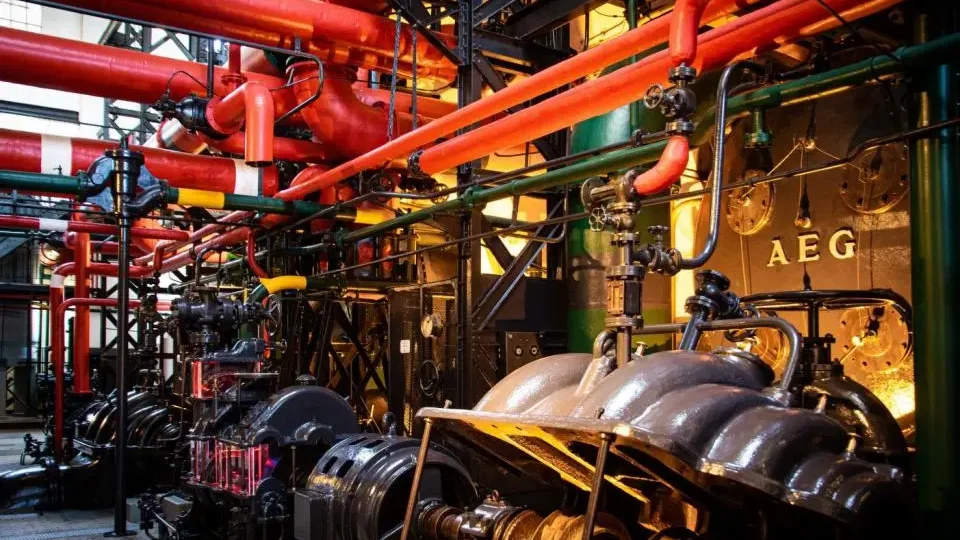 A room brimming with the Industrial Equipment Application, featuring red and green pipes, metal gears, and components, including machines labeled "AEG.