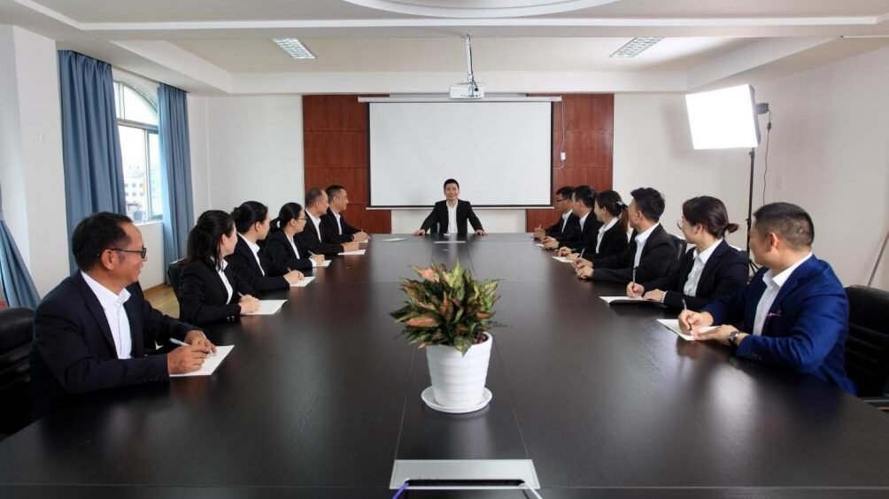 A group of professionals in business attire sit around a long conference table, attentively listening to a person standing at the front near a projector screen, discussing Liangjiang Cabinet Lock Co., Ltd. 1.