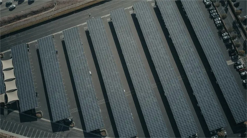 Solar PV Equipment Aerial view of a parking lot featuring multiple rows of carports equipped with Solar PV Equipment. Adjacent to the lot are roadways and several vehicles.
