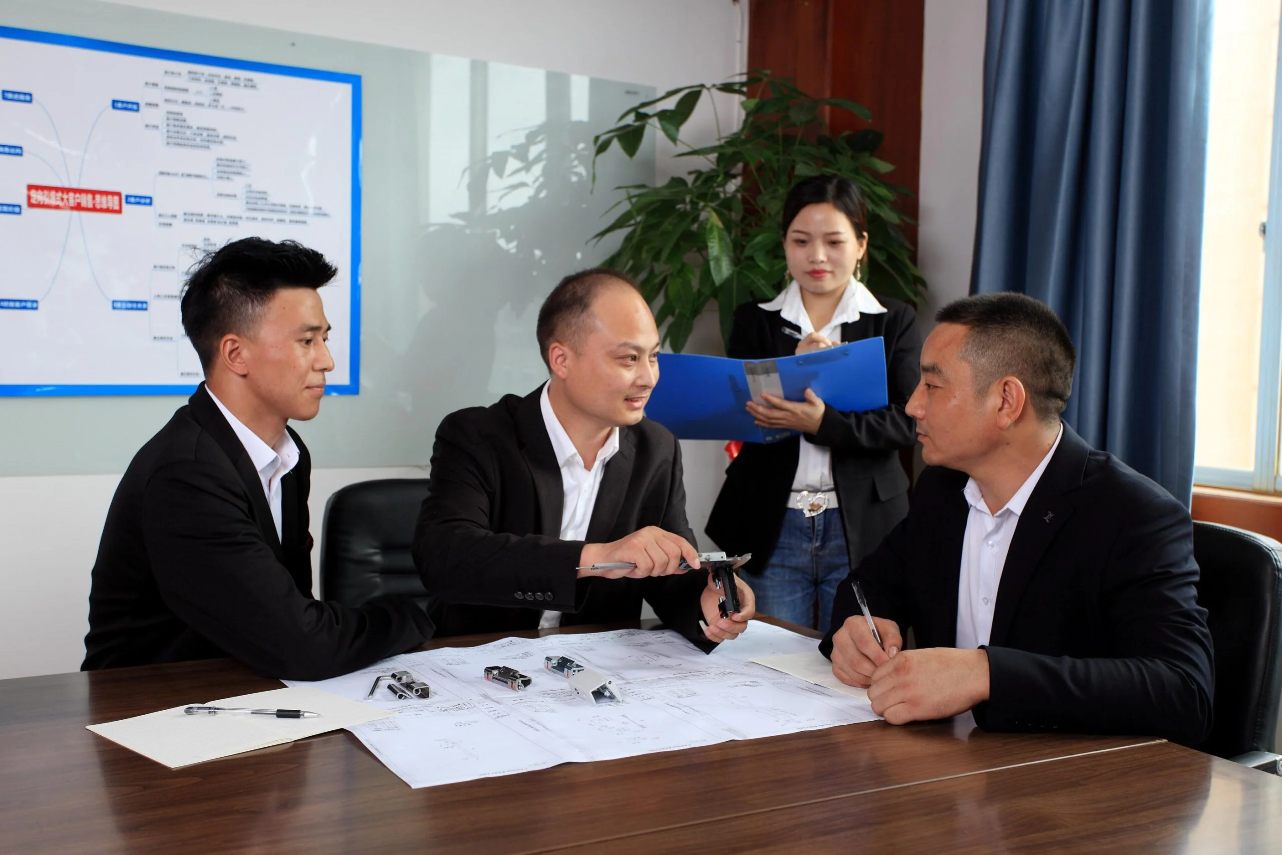 Four people in a meeting room, three seated at a table with documents and one standing holding a blue Brand Concept folder. The seated individuals appear to be discussing a small mechanical part.