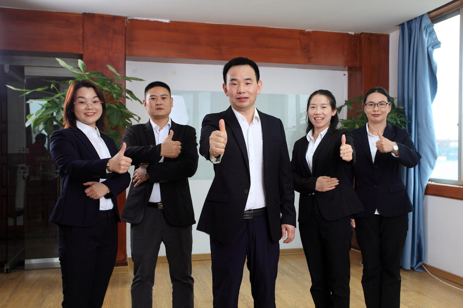 Five business professionals in suits stand indoors, looking at the camera and giving a thumbs up while using Contact.
