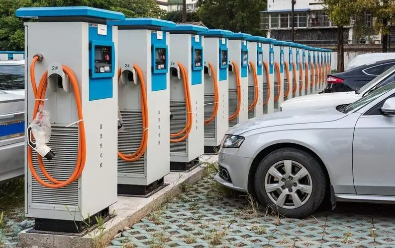 A row of New Energy Vehicle Charging Stations is lined up with orange cables attached, and silver cars are parked next to them.