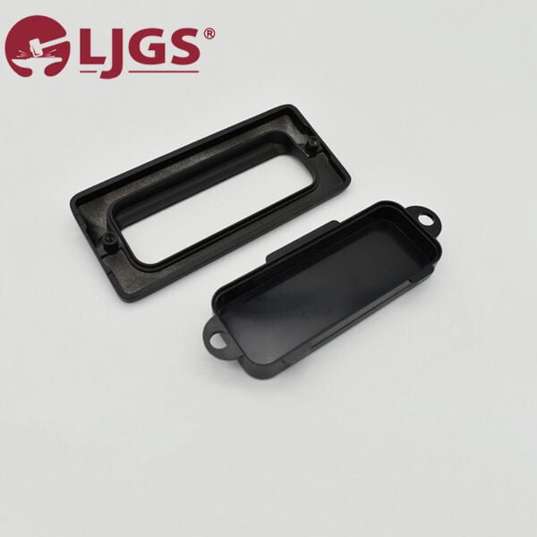 Two black rectangular plastic LS122 (6) components, made from Zinc Alloy, with one having a recessed area and tabs on the sides. They are elegantly placed on a light gray surface. The red LJGS logo is prominently displayed in the top left corner, adding a vibrant touch.