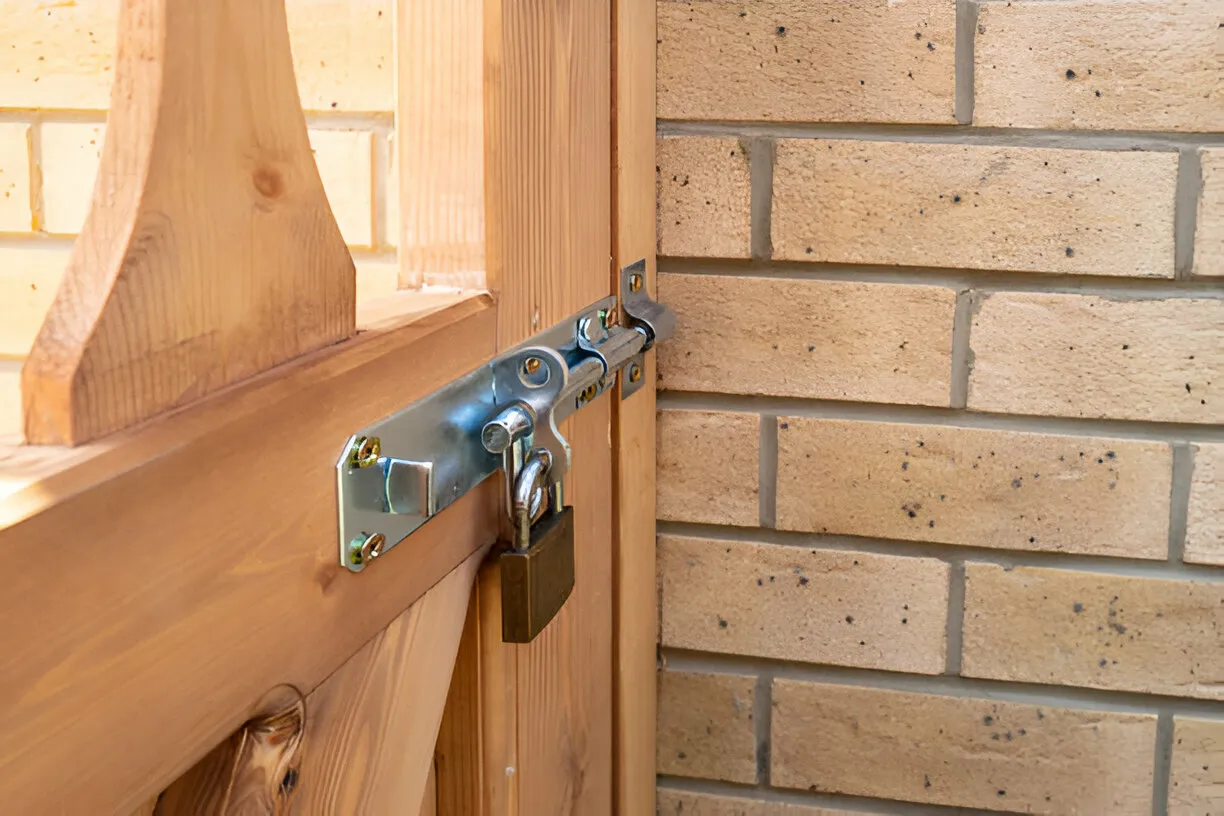 A wooden gate, installed with a tower bolt lock for security, is fastened with a padlock next to a brick wall.